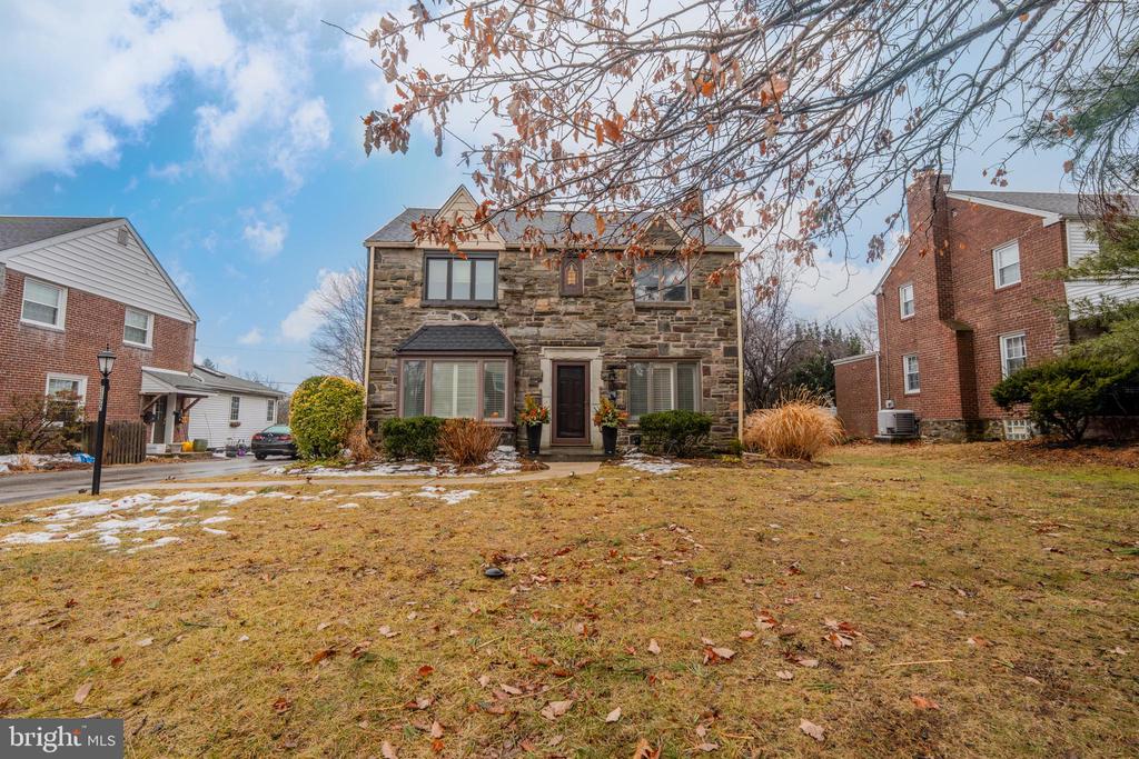 Two-story stone house with a centered entrance, bare branches overhead, and a snow-patched front lawn.