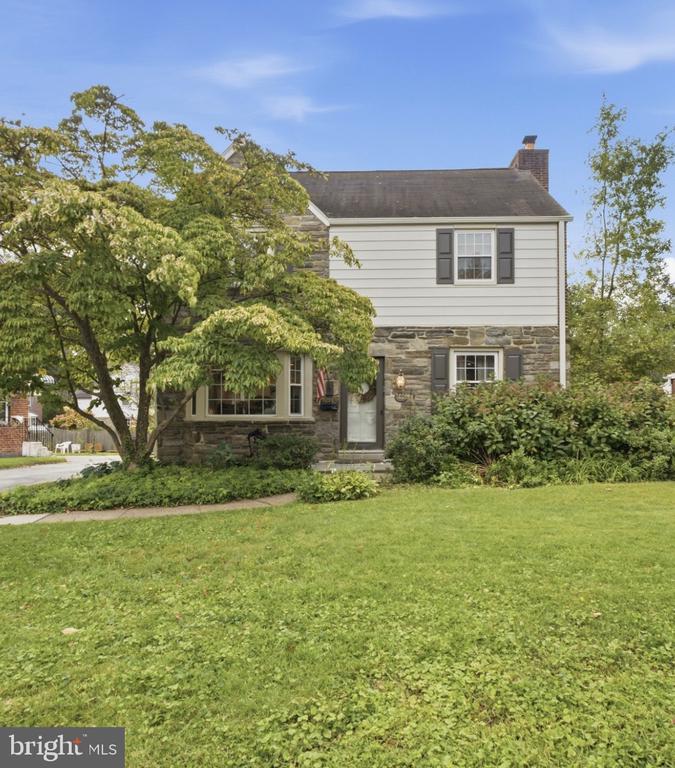 Two-story stone-and-siding house with dark shutters, leafy trees, and a broad front lawn.