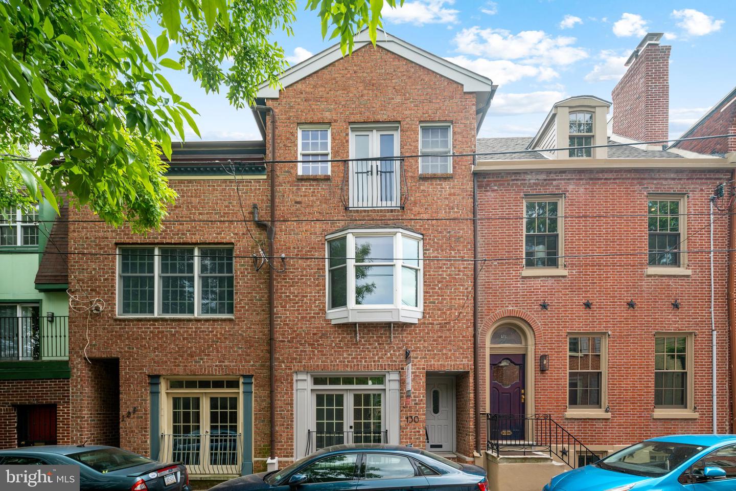 Red-brick rowhouse with a projecting bay window and small upper balcony beneath leafy branches.