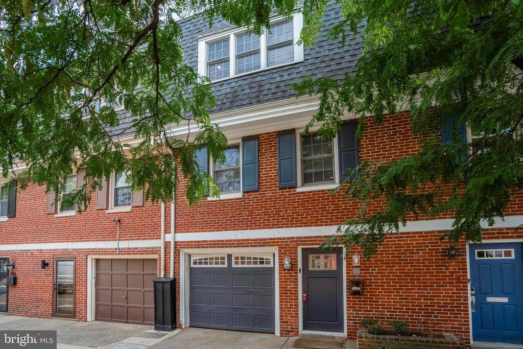 Red-brick townhouse with blue-gray shutters and garage doors beneath leafy trees.