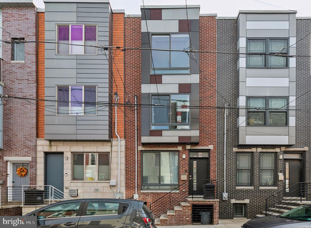Attached brick and gray-clad rowhomes with black railings, overhead wires, and parked cars.