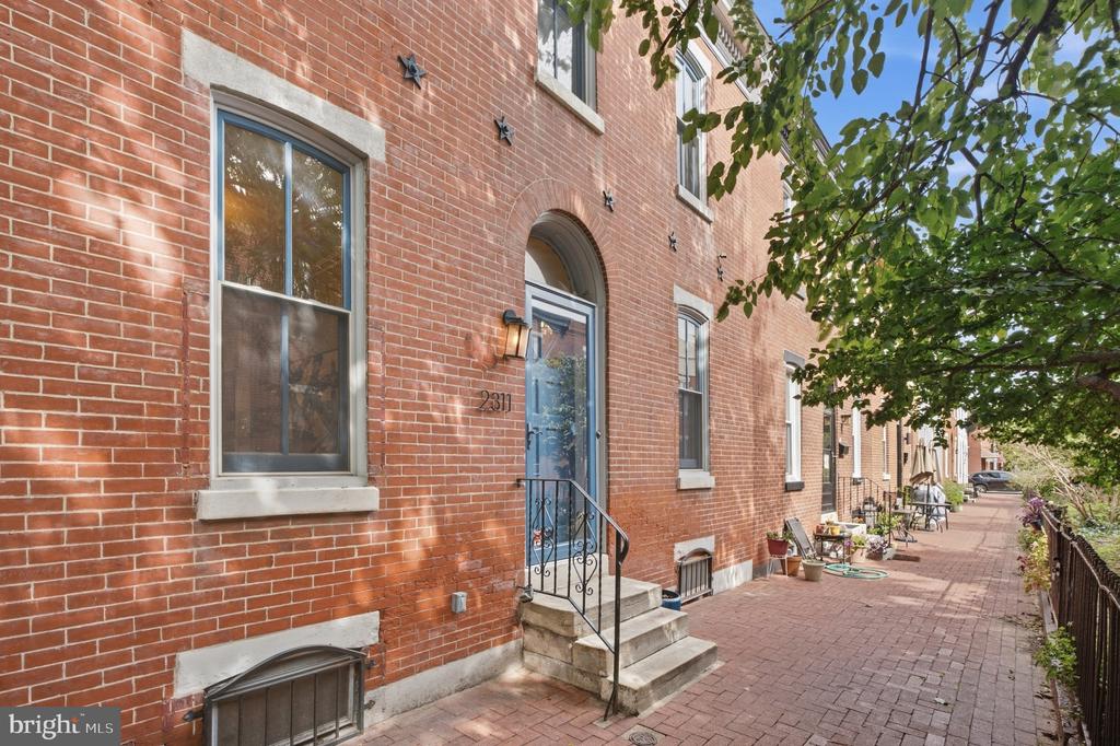 Red-brick rowhouse with an arched blue doorway and leafy shade over the brick sidewalk.
