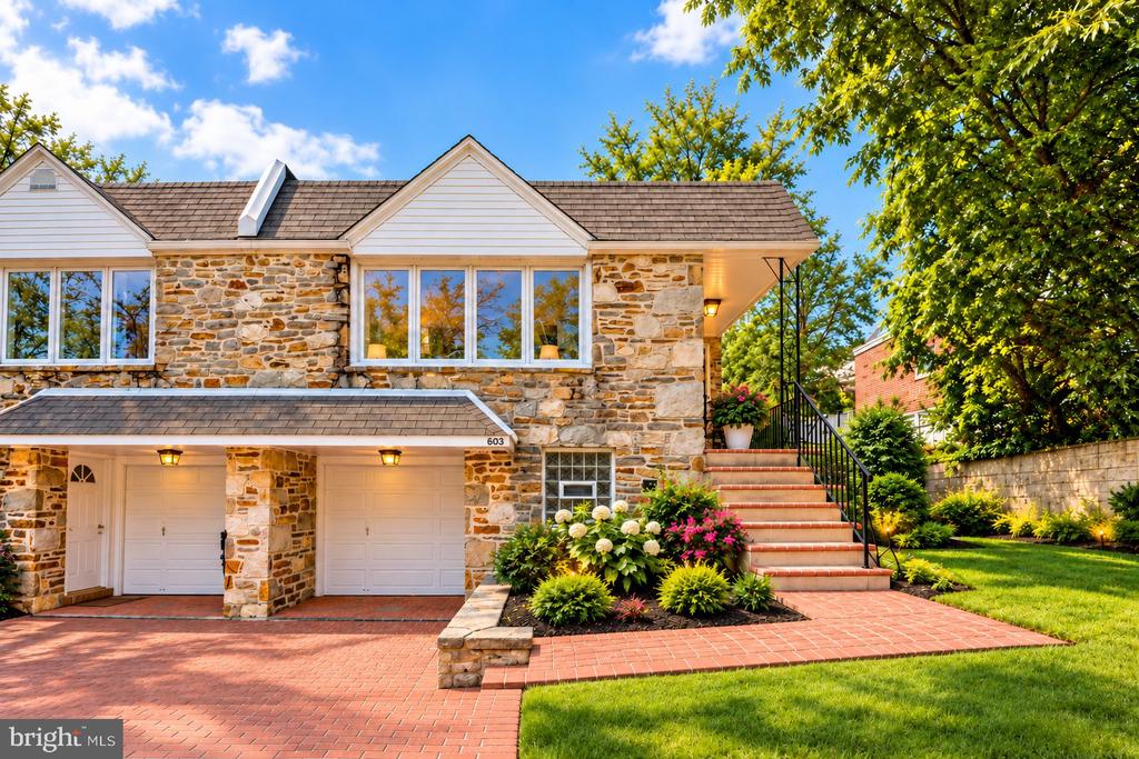 Stone-front house with two garage doors, a brick driveway, and landscaped steps to the entrance.
