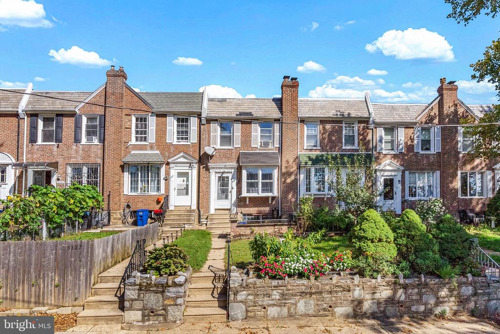 Brick rowhouses with front gardens and stone retaining walls under a blue sky.