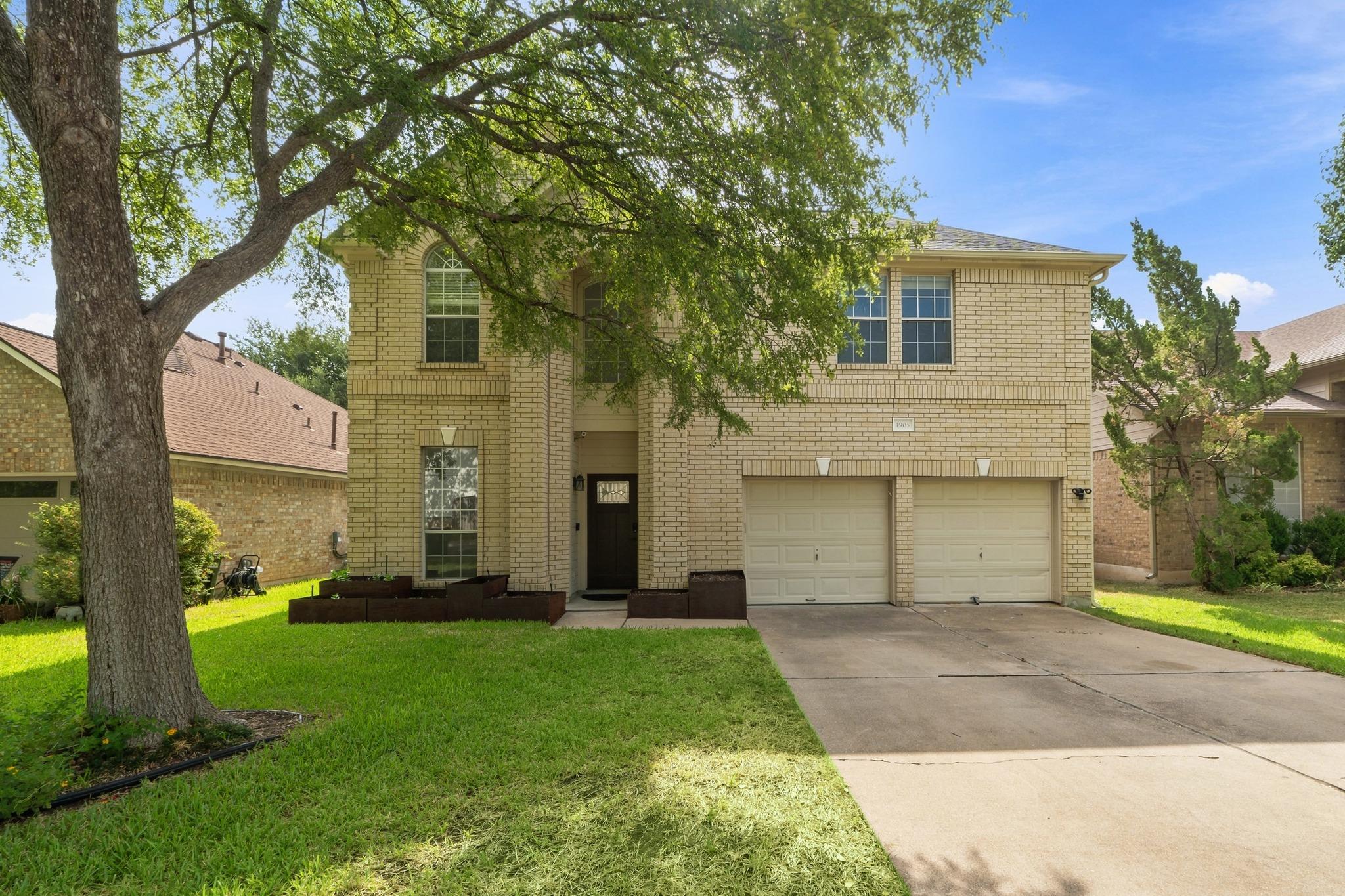 Two-story tan brick house with a double garage, shaded by a large tree over the front lawn.