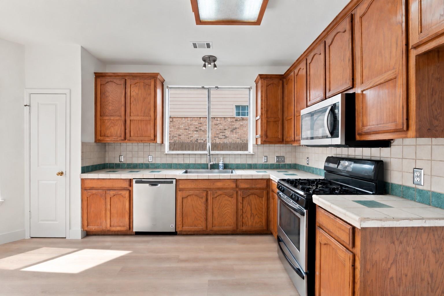 Kitchen with warm wood cabinetry, tiled counters, and stainless steel appliances.