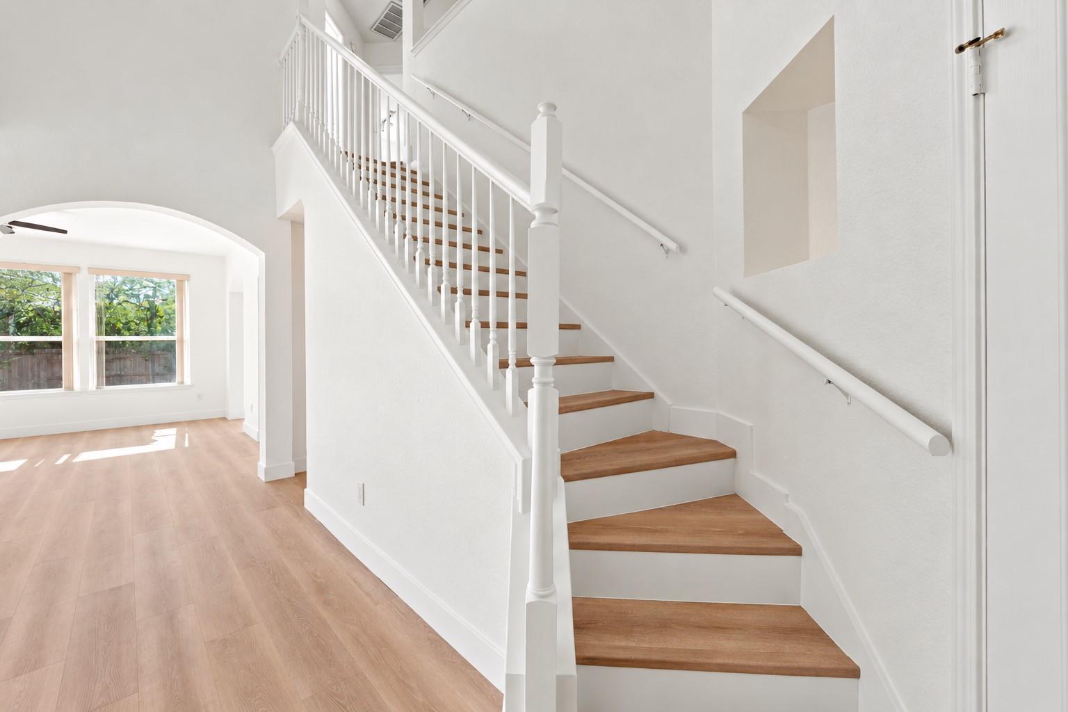 White staircase with wood treads beside a sunlit living area with an arched window alcove.