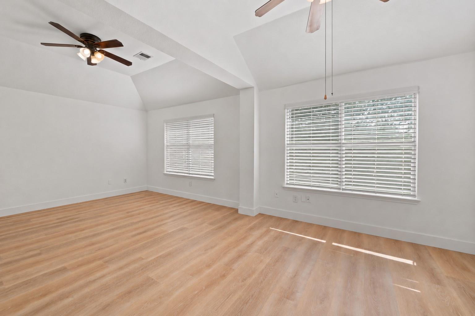 Empty room with light wood flooring, vaulted ceiling, ceiling fans, and windows with white blinds.