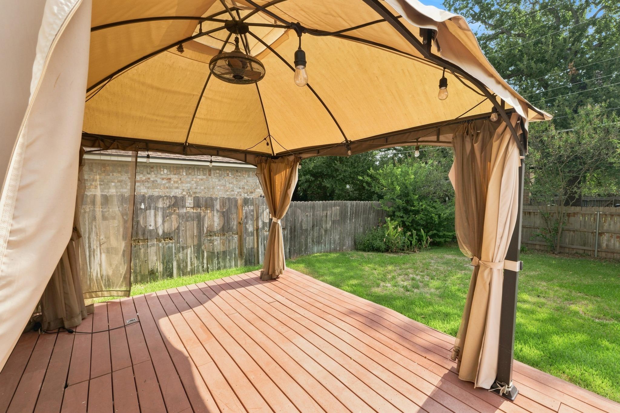 Wooden backyard deck under a curtained gazebo, overlooking a fenced lawn.