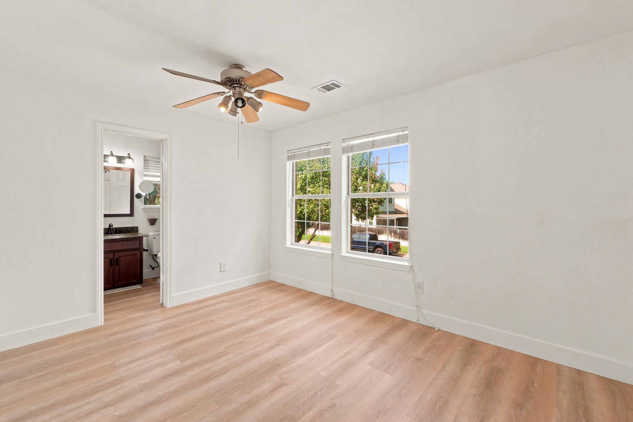 Empty bedroom with light wood-look flooring, a ceiling fan, two windows, and a bathroom doorway.