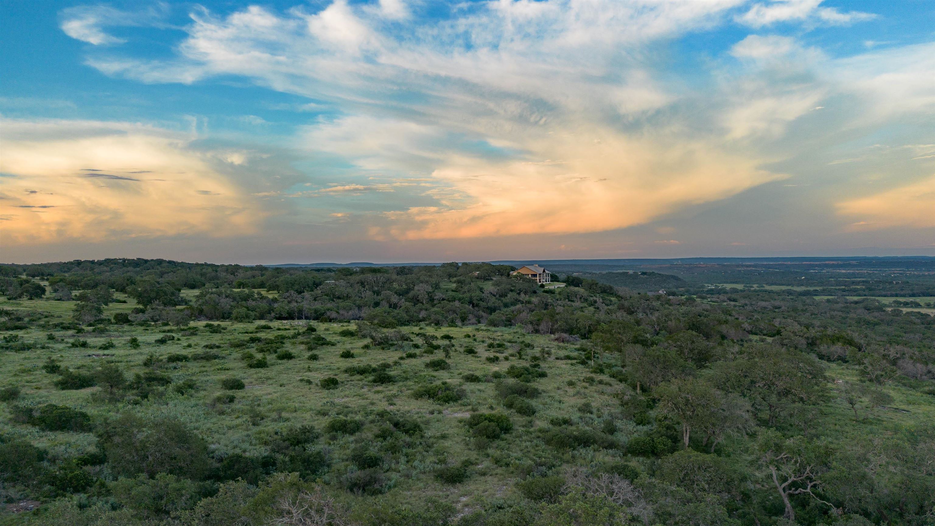 Aerial view of a brush-covered tract with scattered trees and a distant house beneath a sunset sky.