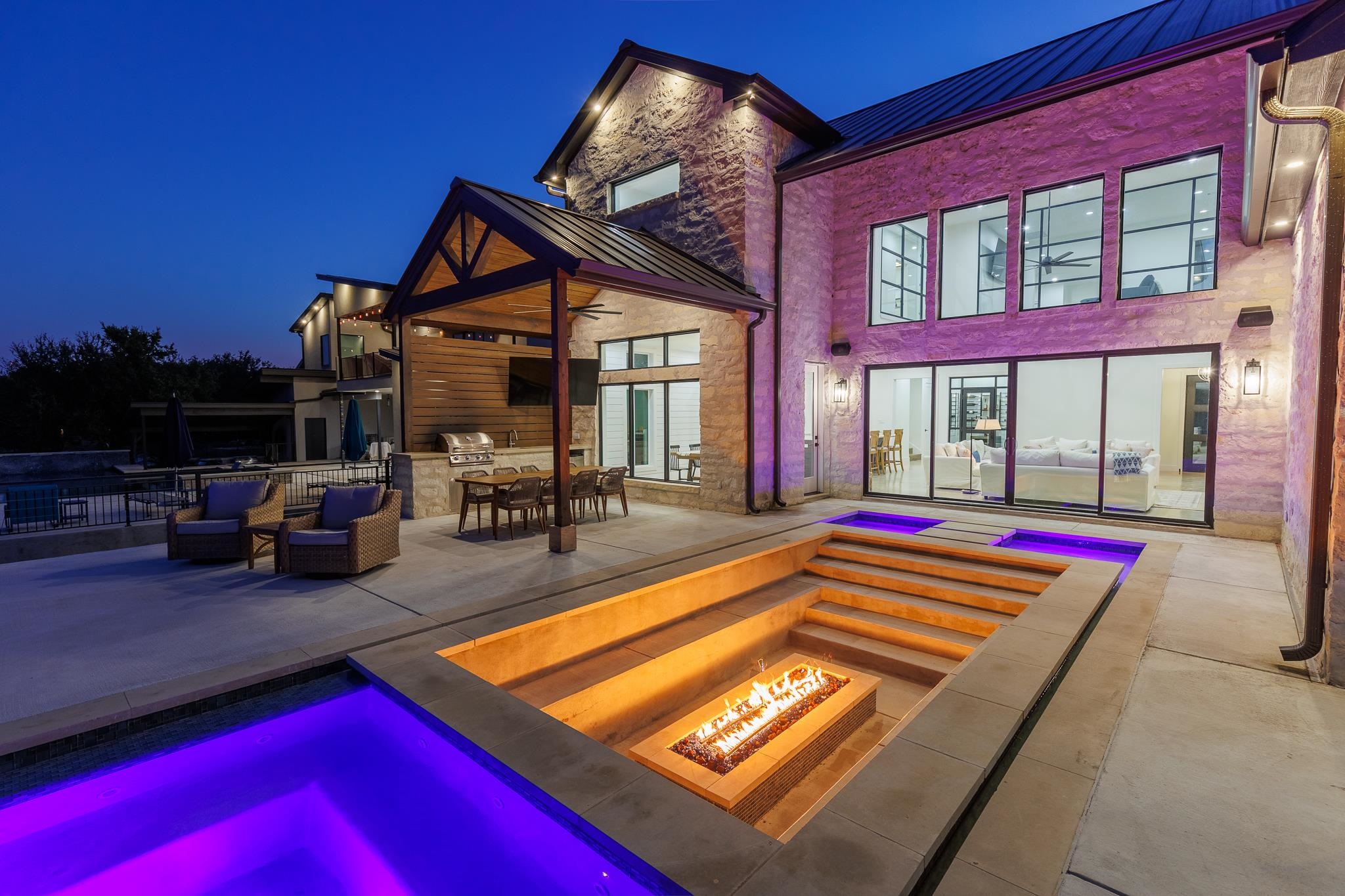 Modern stone home beside an illuminated pool and sunken fire-pit patio at night.