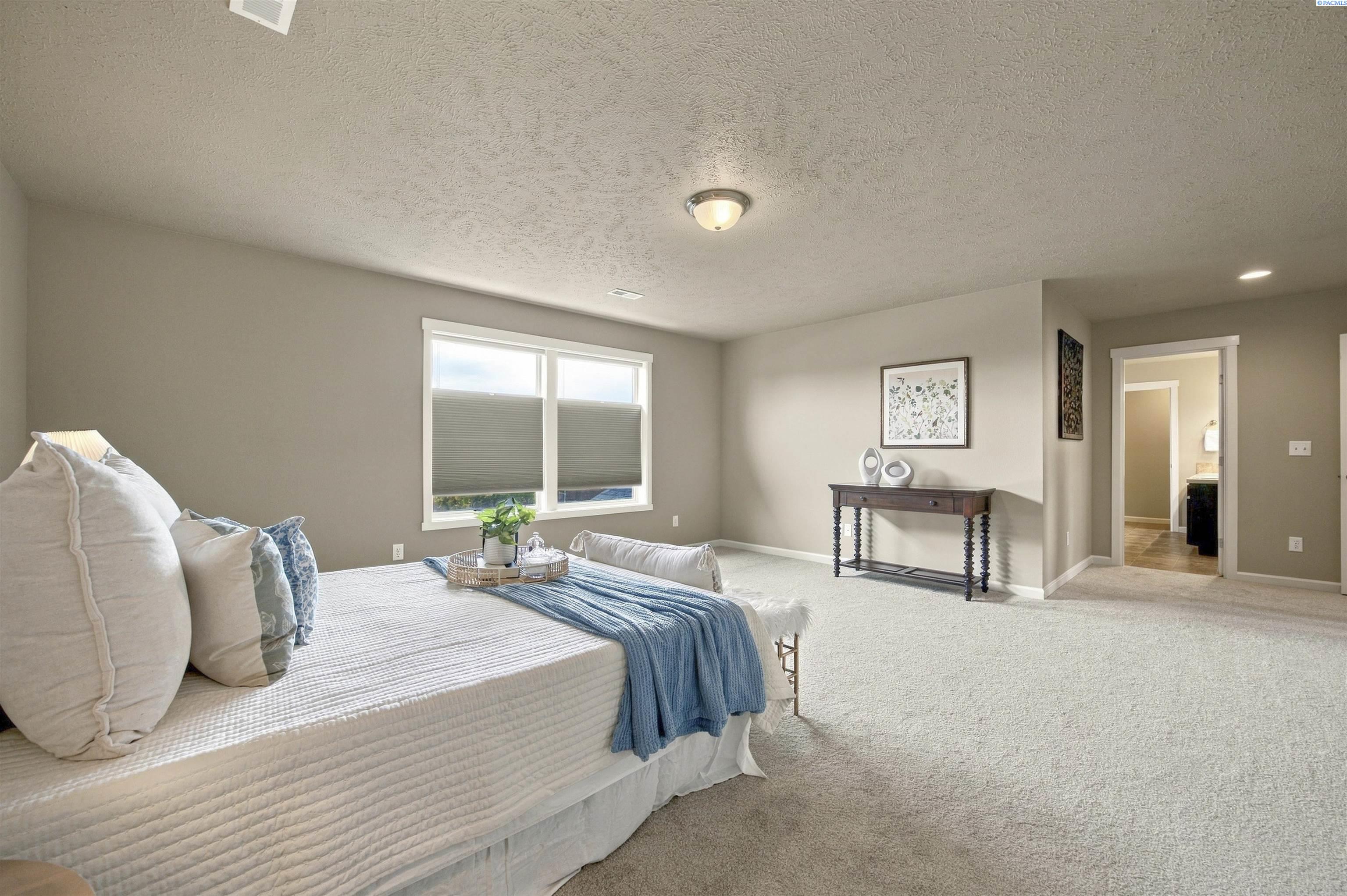 Staged bedroom with a white bed, blue throw, carpet, and doorway to a bathroom.