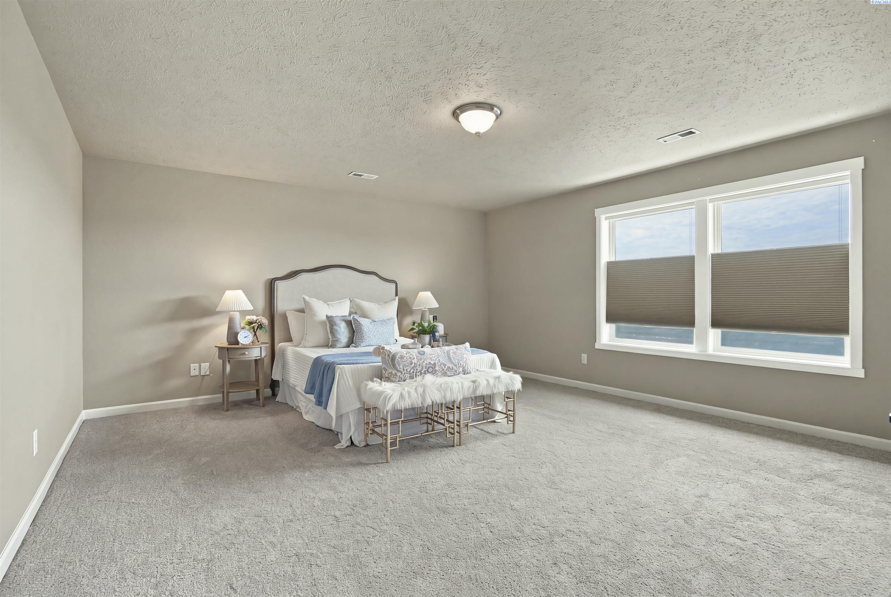 Spacious carpeted bedroom with a staged bed and two large windows with partially lowered shades.