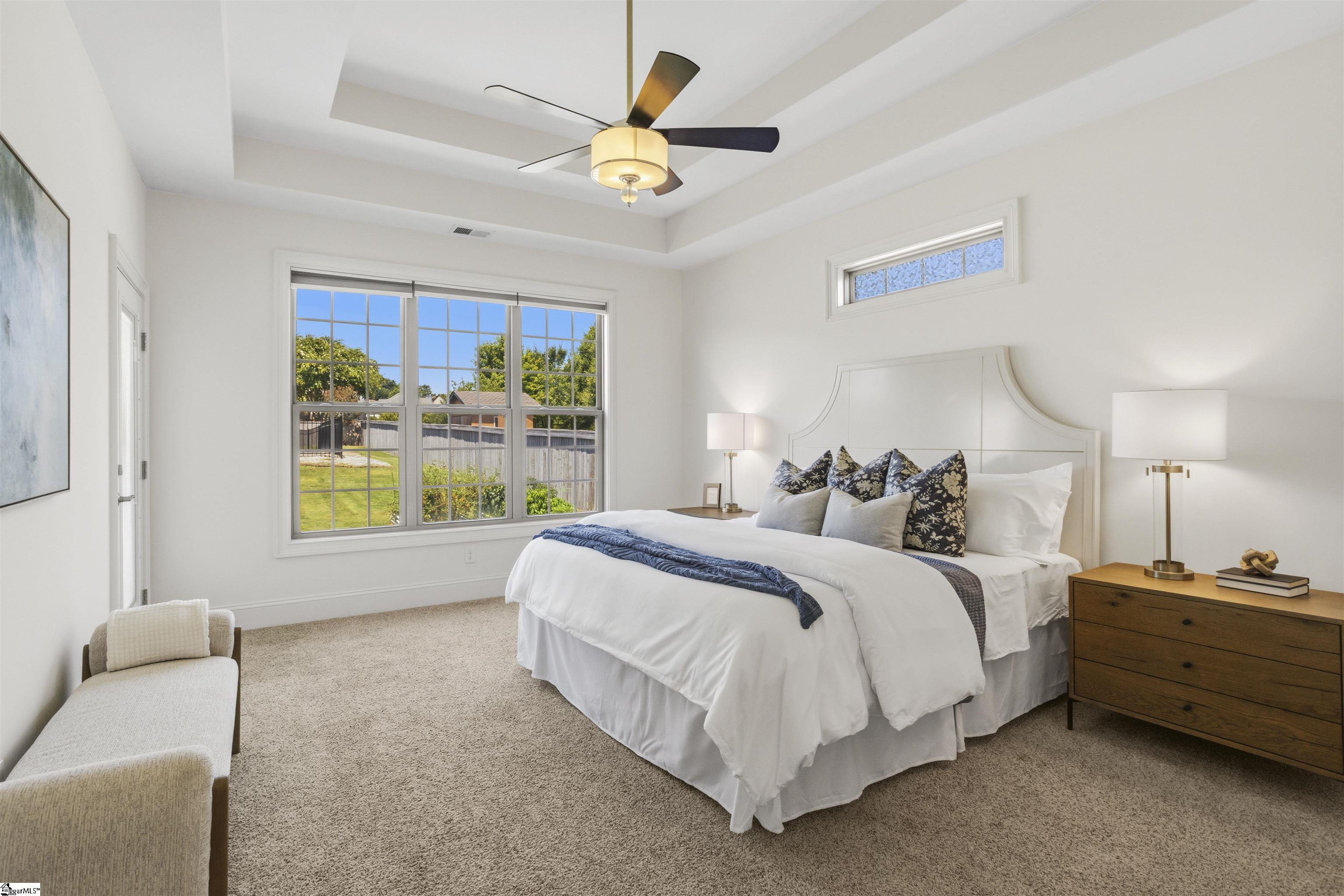 Staged bedroom with a white bed, blue pillows, tray ceiling, and large window overlooking a fenced yard.