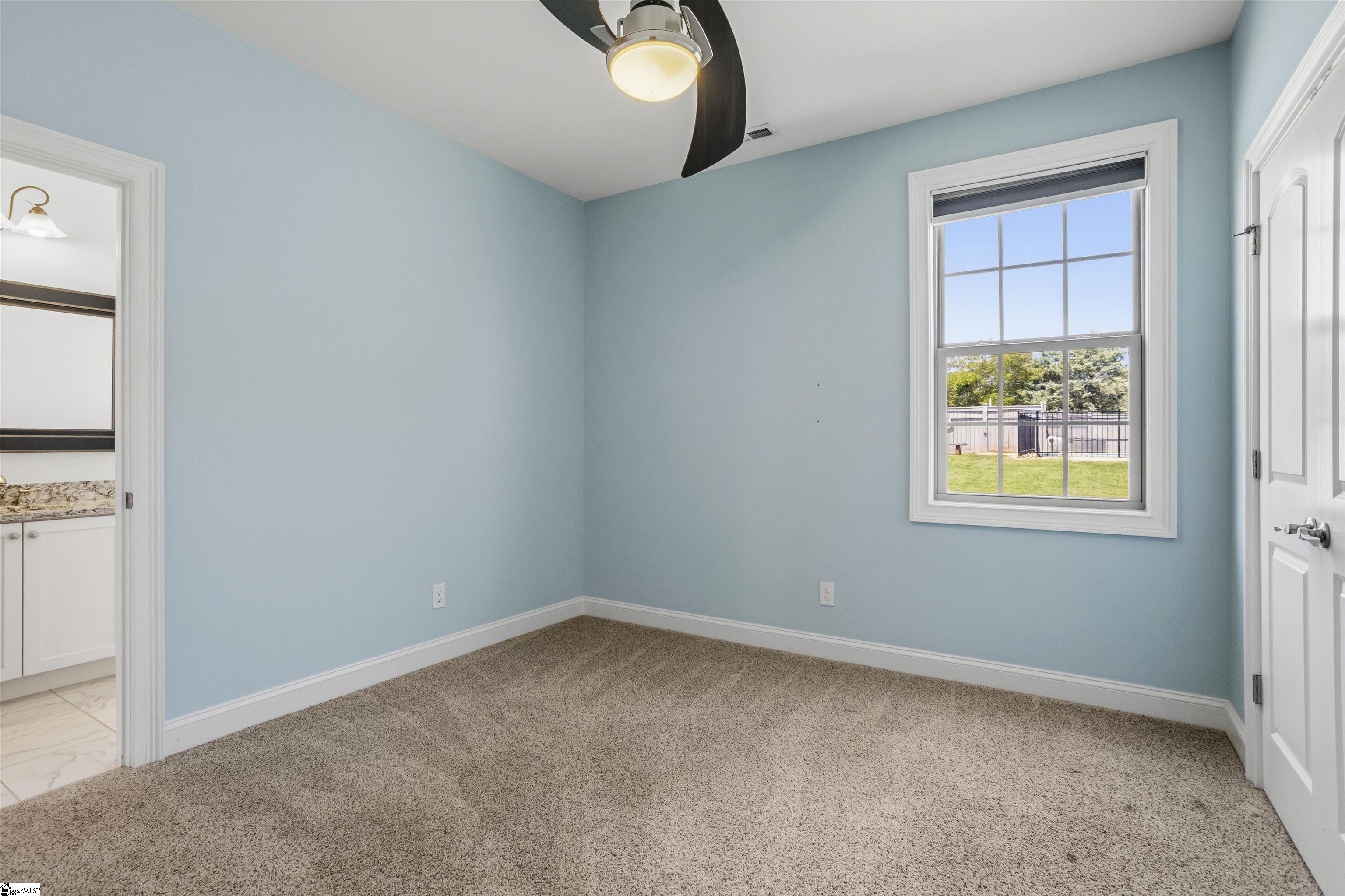 Empty blue bedroom with beige carpet, ceiling fan, and a window overlooking the backyard.
