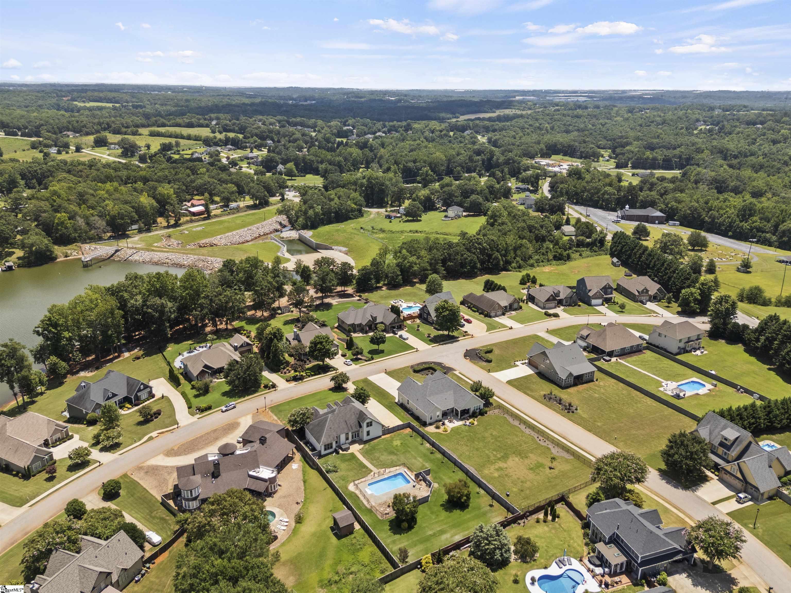 Aerial view of homes with fenced yards and pools beside a pond and tree-lined streets.