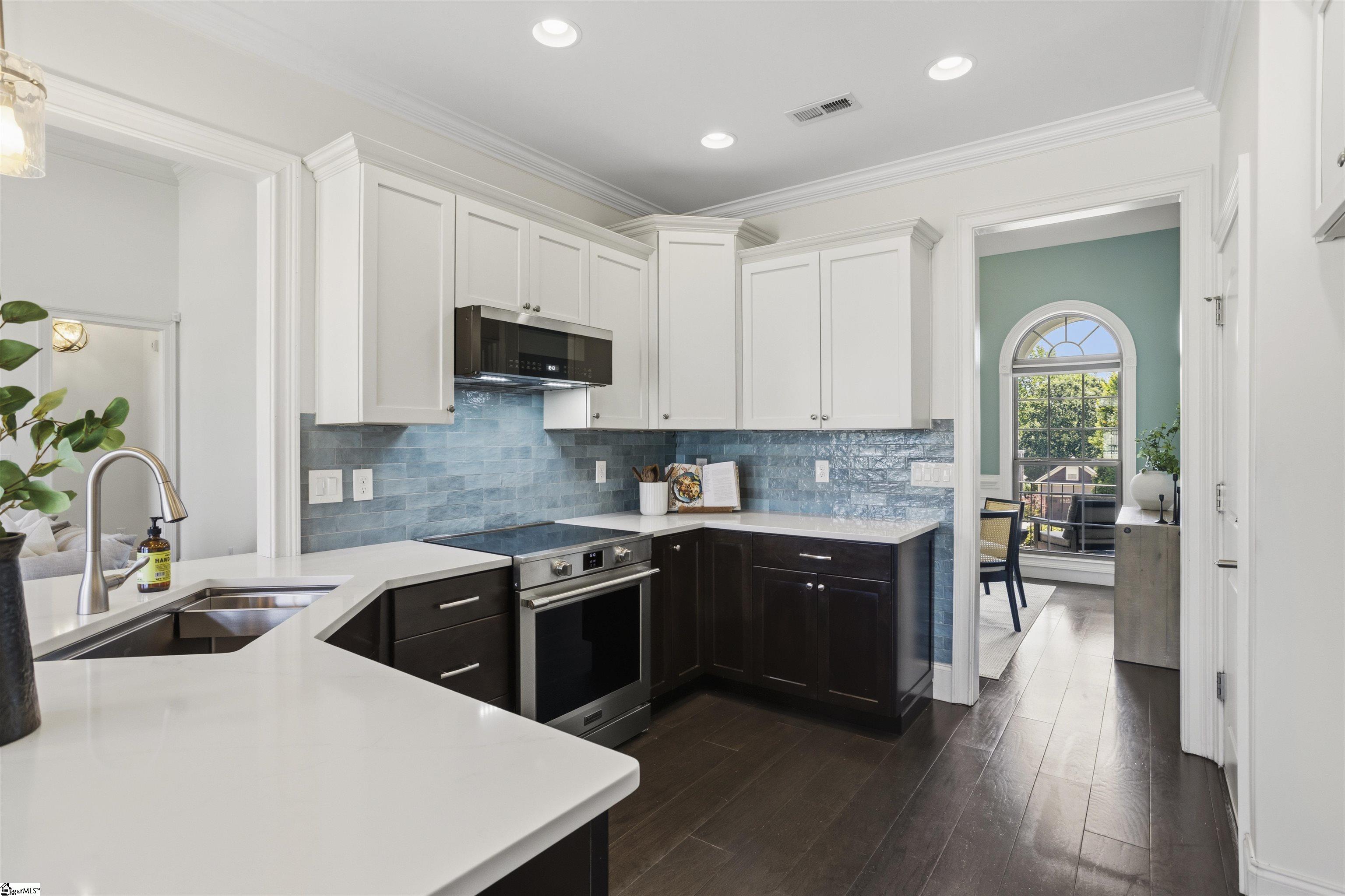 Kitchen with blue tile backsplash, white counters, dark cabinets, and an adjoining room with an arched window.