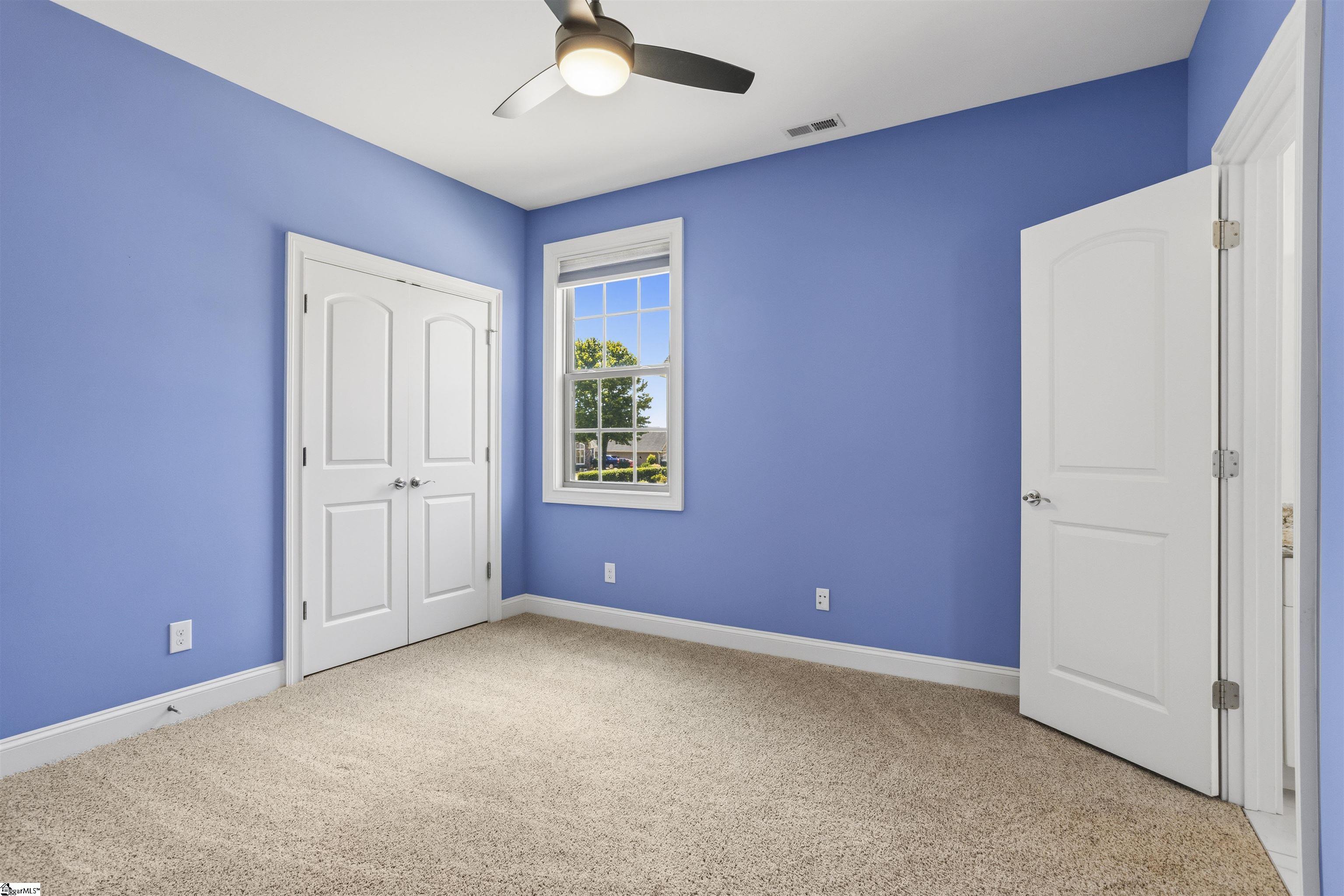 Empty bedroom with blue walls, beige carpet, white closet doors, and a ceiling fan.