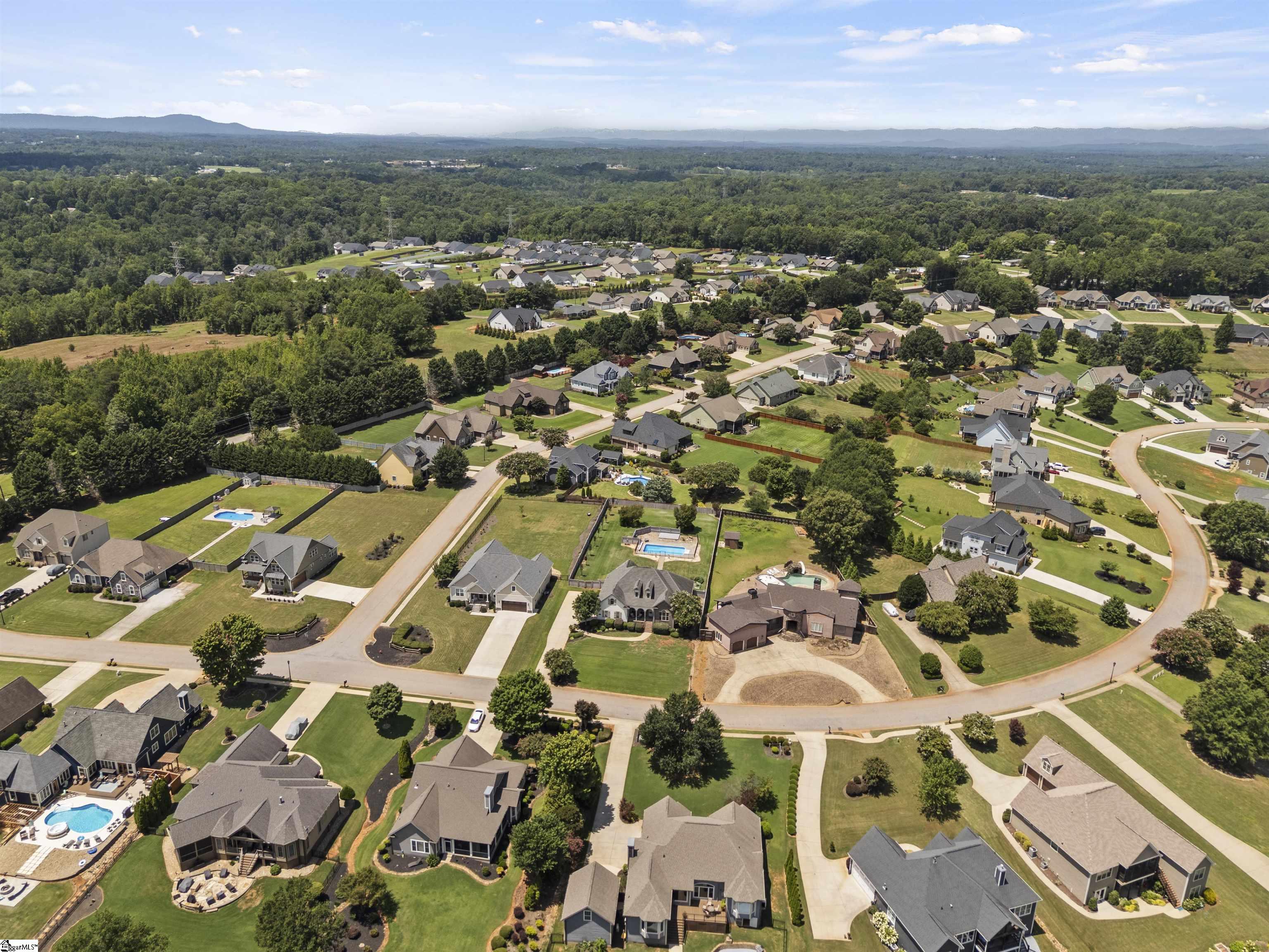Aerial view of a suburban neighborhood with wooded surroundings, backyard pools, and a distant ridge.