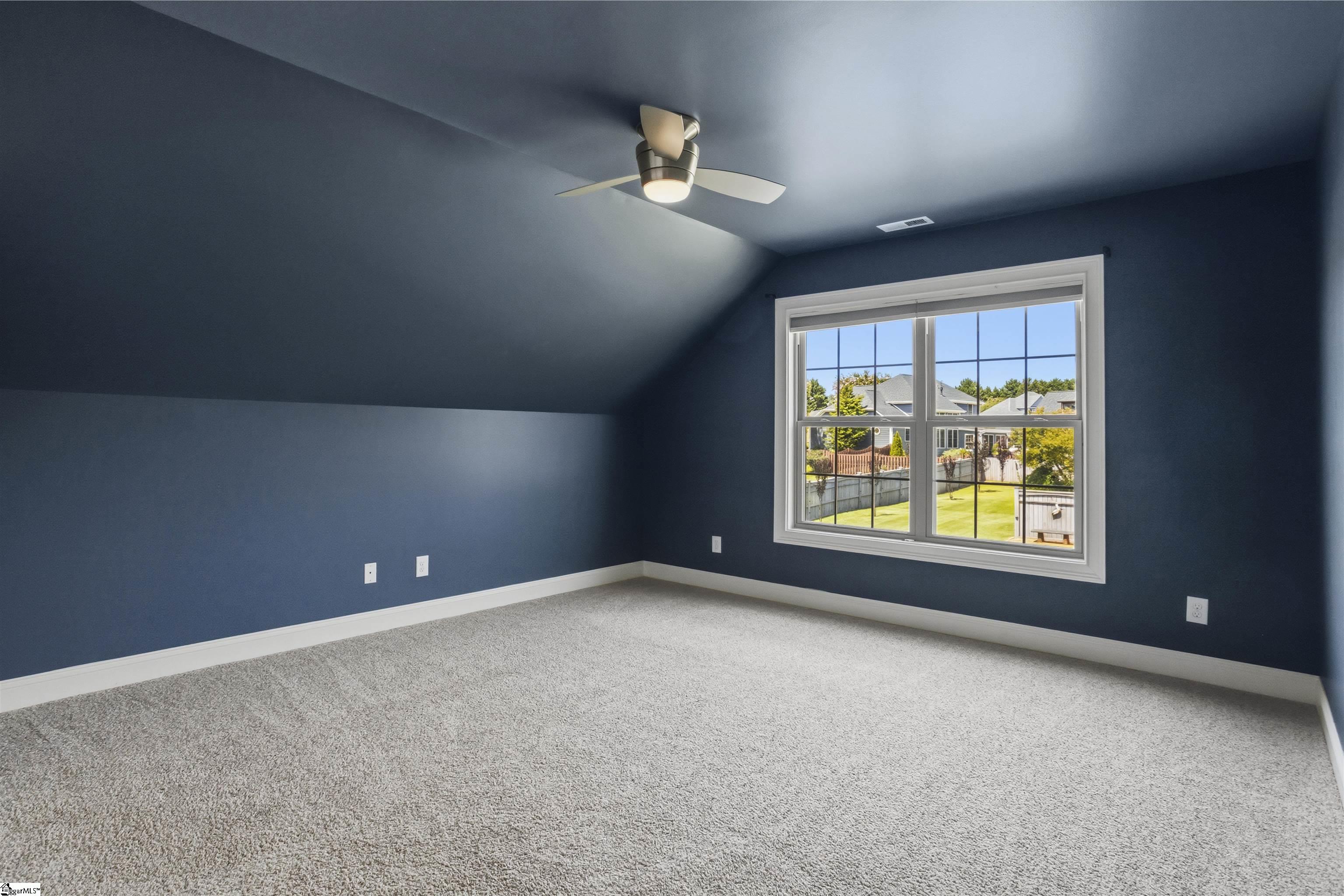 Empty blue room with a sloped ceiling, gray carpet, ceiling fan, and window overlooking a fenced yard.