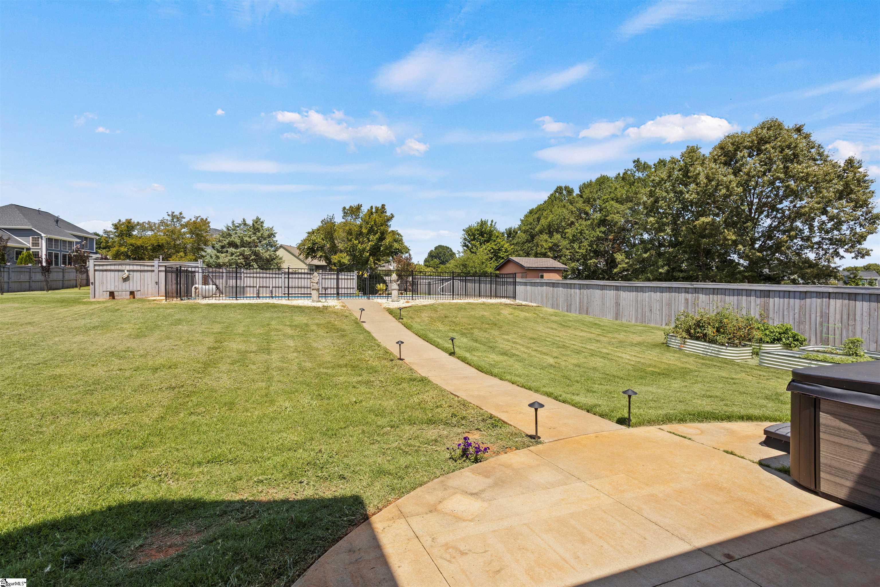 Fenced backyard with a large lawn, patio walkway, gated pool, raised garden beds, and a hot tub.