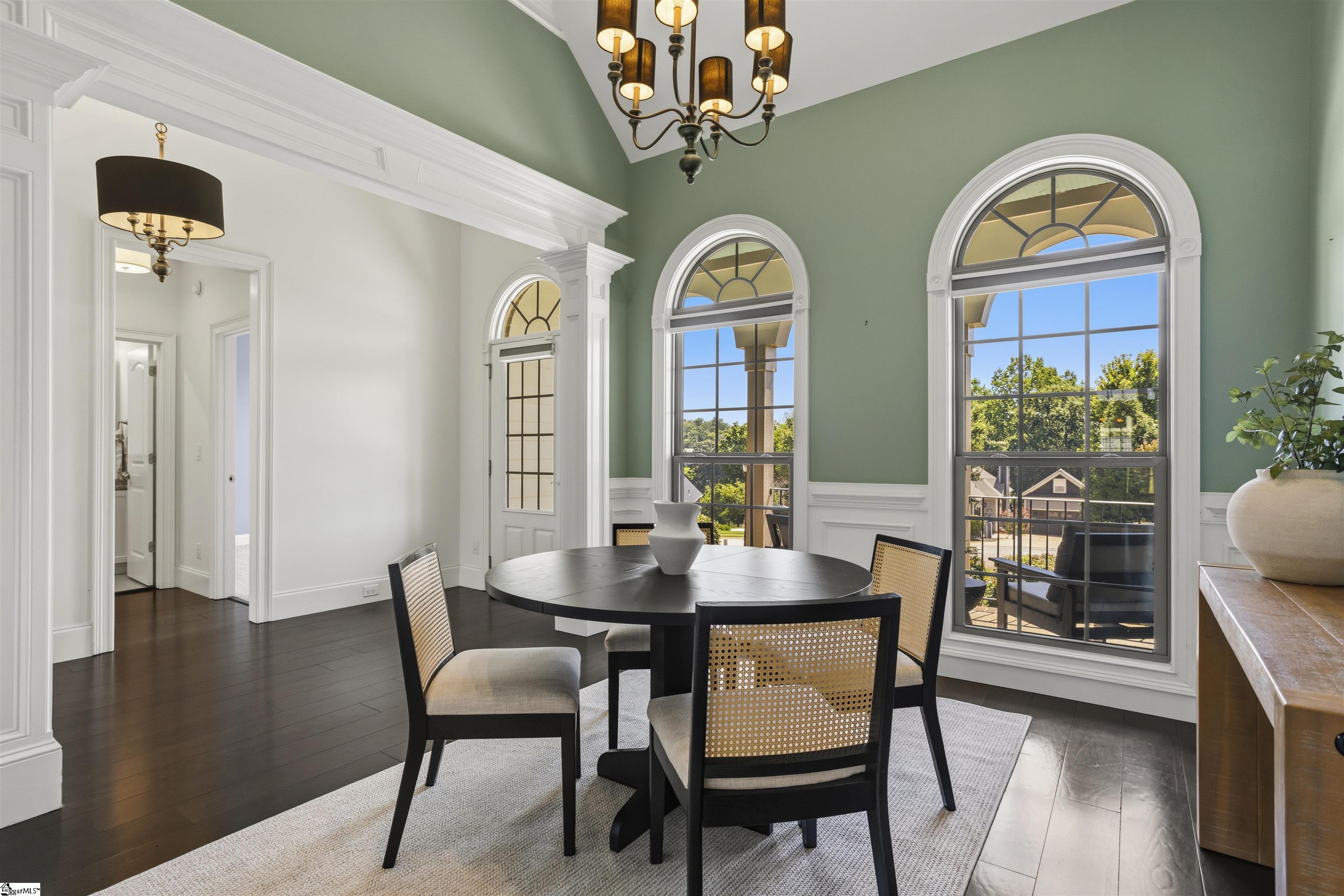 Dining room with a round black table, sage-green walls, and three tall arched windows.