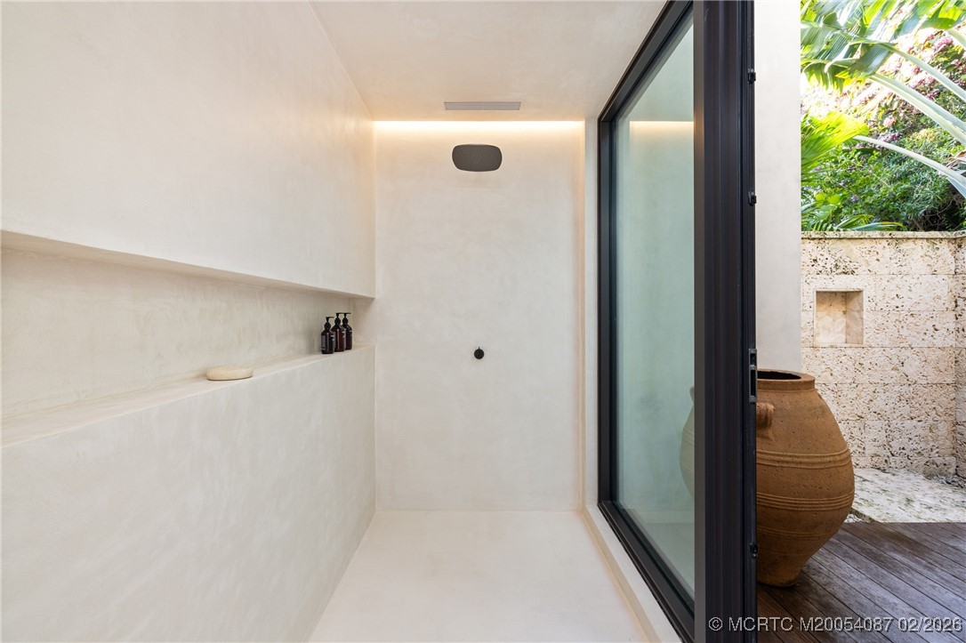 White stone shower with a built-in shelf and black-framed glass opening to a tropical courtyard.