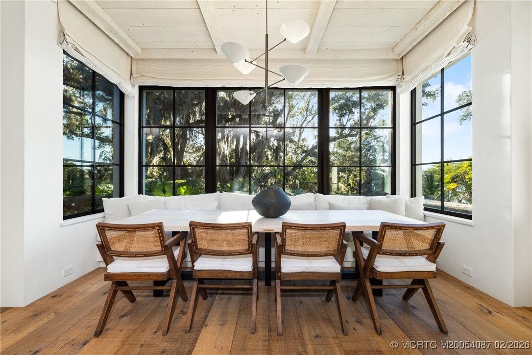 Dining room with woven wooden chairs, a white table, and broad windows overlooking leafy trees.