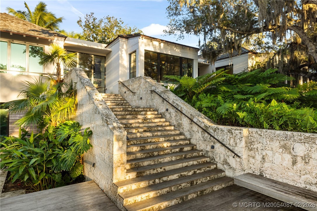 Modern white home beyond a broad stone staircase bordered by tropical plants and mature trees.