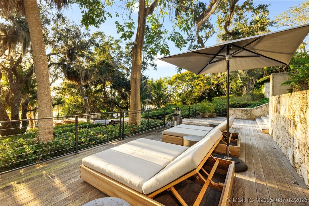 Wooden deck with cushioned loungers beneath a large umbrella, surrounded by mature trees.