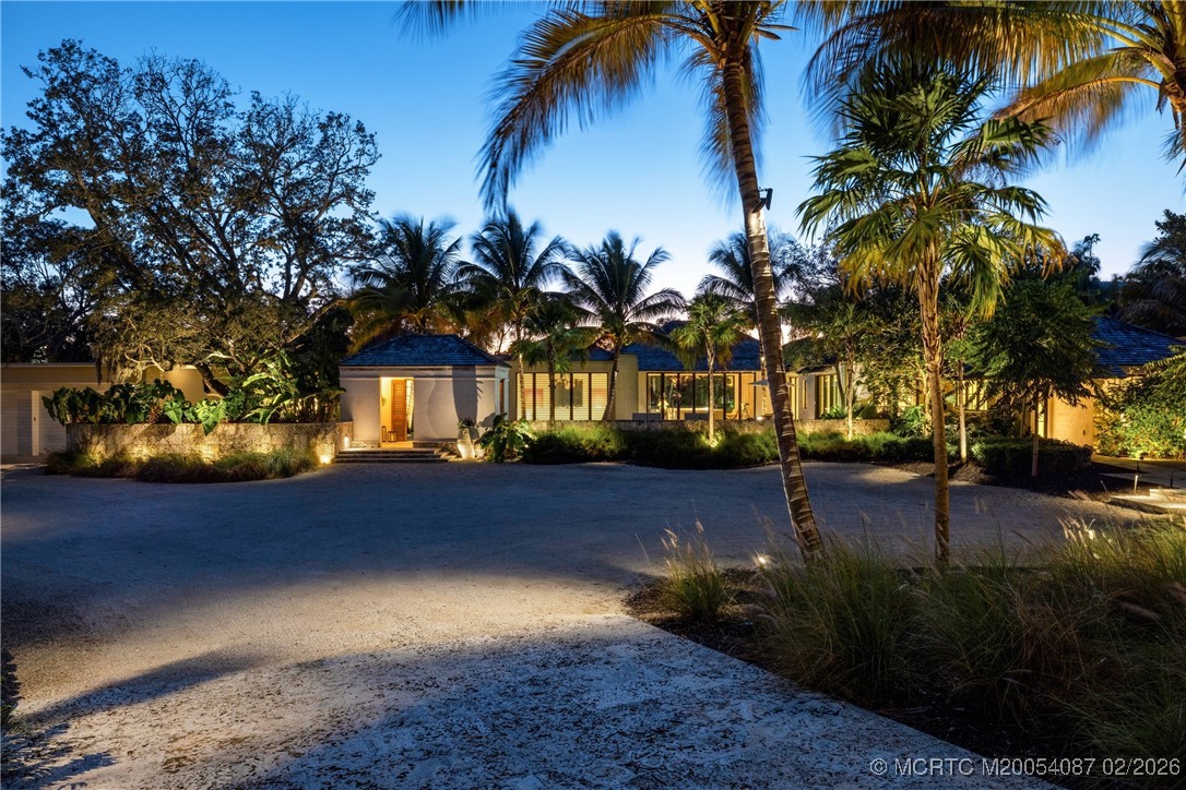 Single-story home with an illuminated entrance, palm trees, and a broad gravel driveway at dusk.