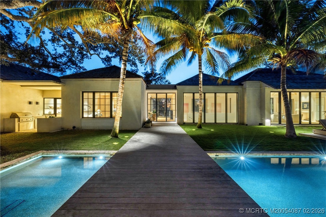 Modern home at dusk, framed by palm trees, with a wooden walkway between two pool sections.