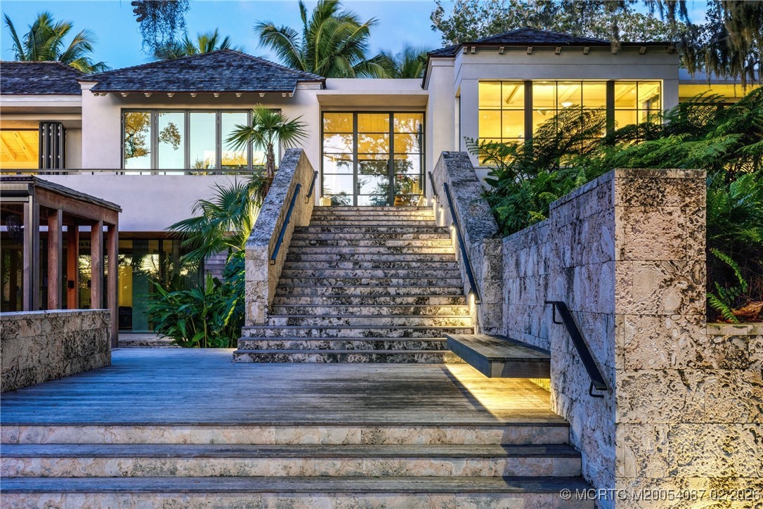 Modern tropical home with a broad stone entrance staircase, glowing glass windows, and lush palms.