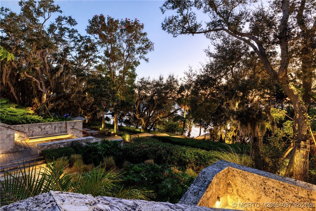 Waterfront garden with illuminated stone terraces, lush tropical landscaping, and mature trees at dusk.