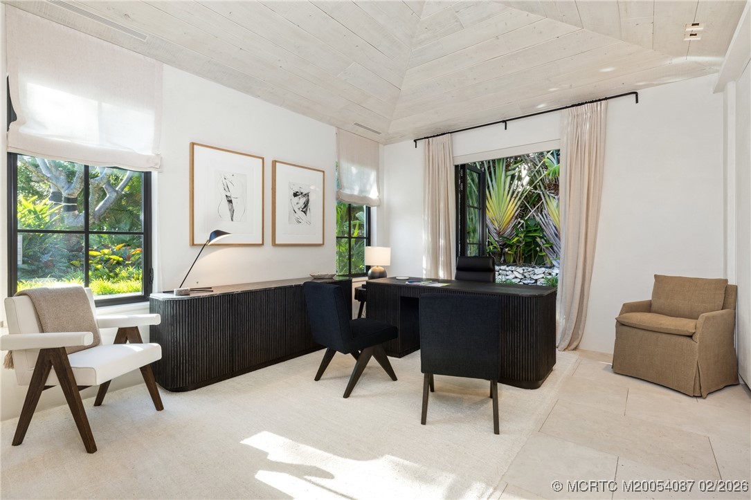 Furnished home office with a dark wood desk, vaulted white ceiling, and windows overlooking tropical greenery.