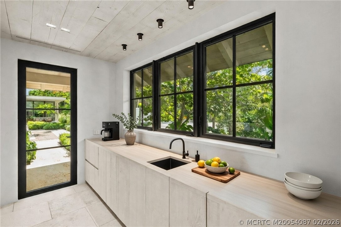 Modern kitchen with pale wood cabinetry, black-framed windows, and a view of lush greenery.