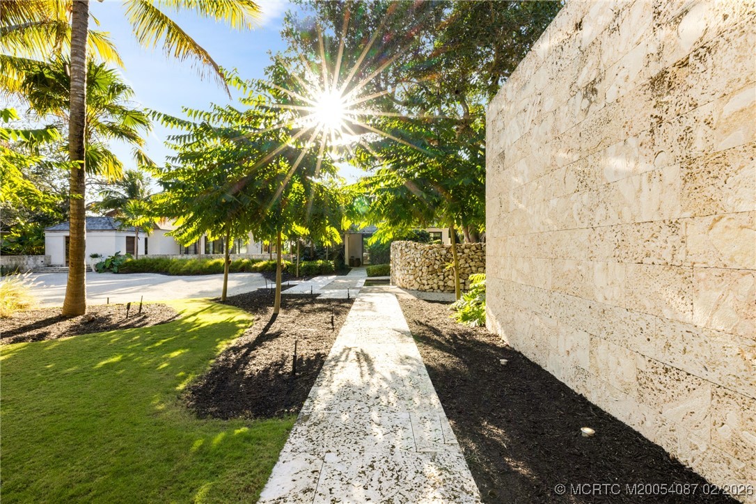 Stone walkway leads toward a home beside a textured wall, palms, and landscaped lawn.