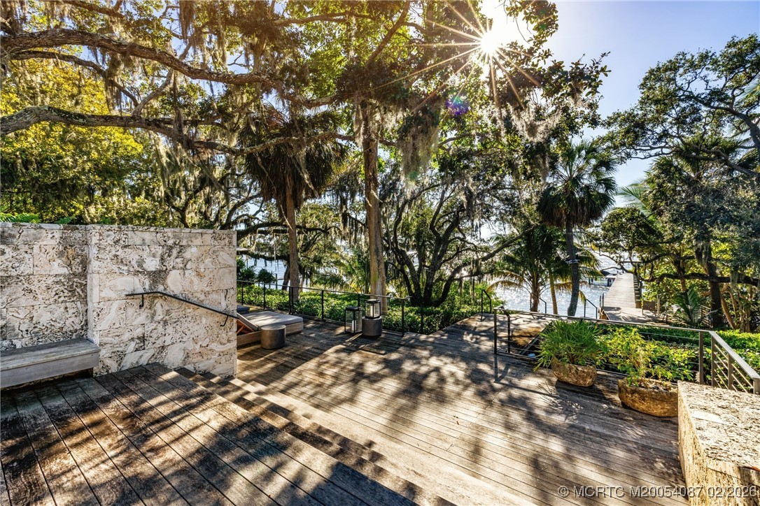 Wooden waterfront deck beneath shady oaks, with stone walls, outdoor seating, and a dock beyond.