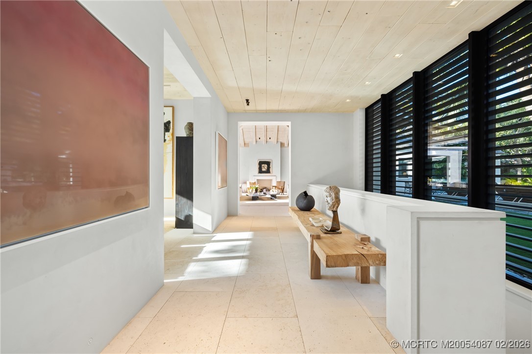 Modern hallway with pale stone floors, black louvered windows, and a wooden bench with sculptural decor.
