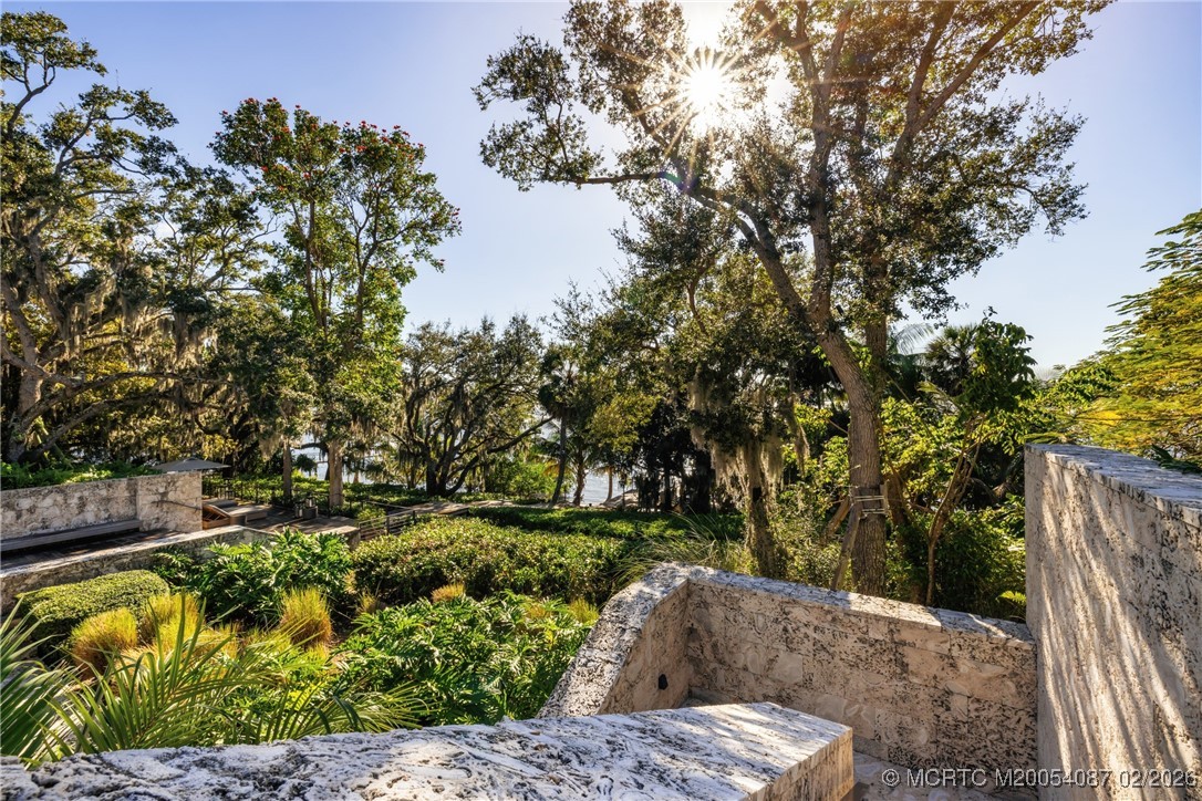 Landscaped stone terrace beneath mature oaks, with tropical plants and water visible beyond.