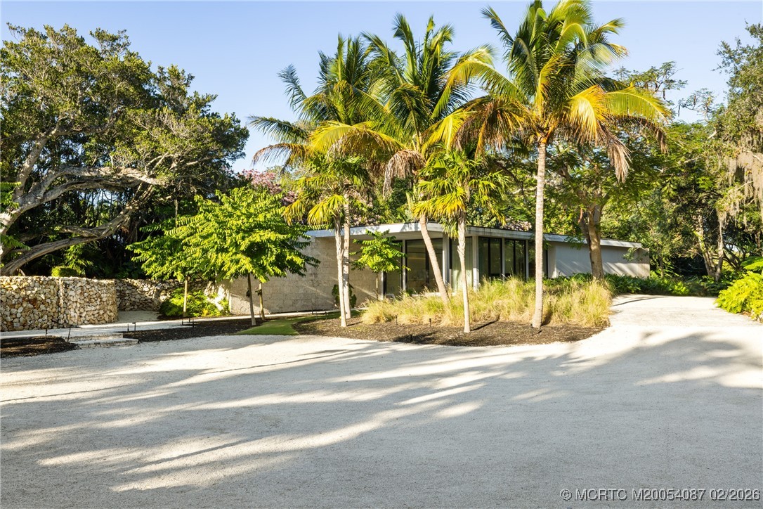 Modern low-profile home behind palm trees, with a broad gravel driveway and low stone wall.