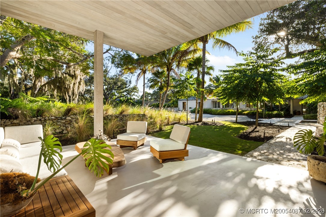 Covered patio with cream lounge chairs and a wood table overlooking a palm-lined garden.