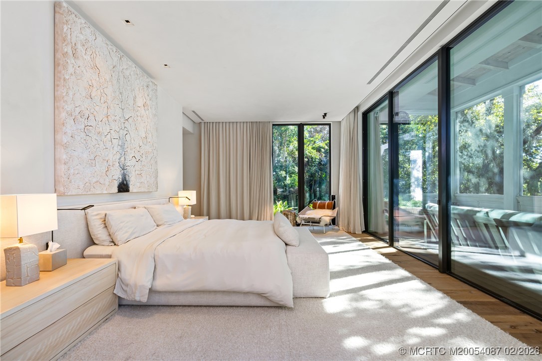 Modern bedroom with a white bed, textured wall art, and floor-to-ceiling glass doors overlooking greenery.