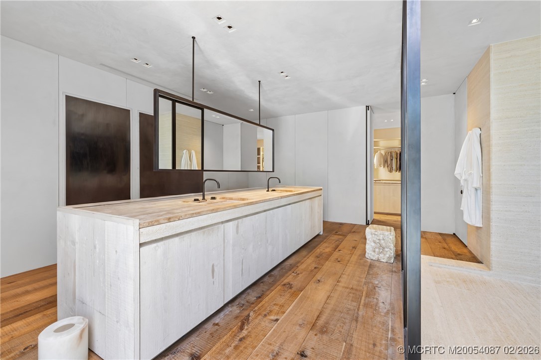 Modern bathroom with a long double vanity, suspended mirror, warm wood floors, and an open walk-in closet.