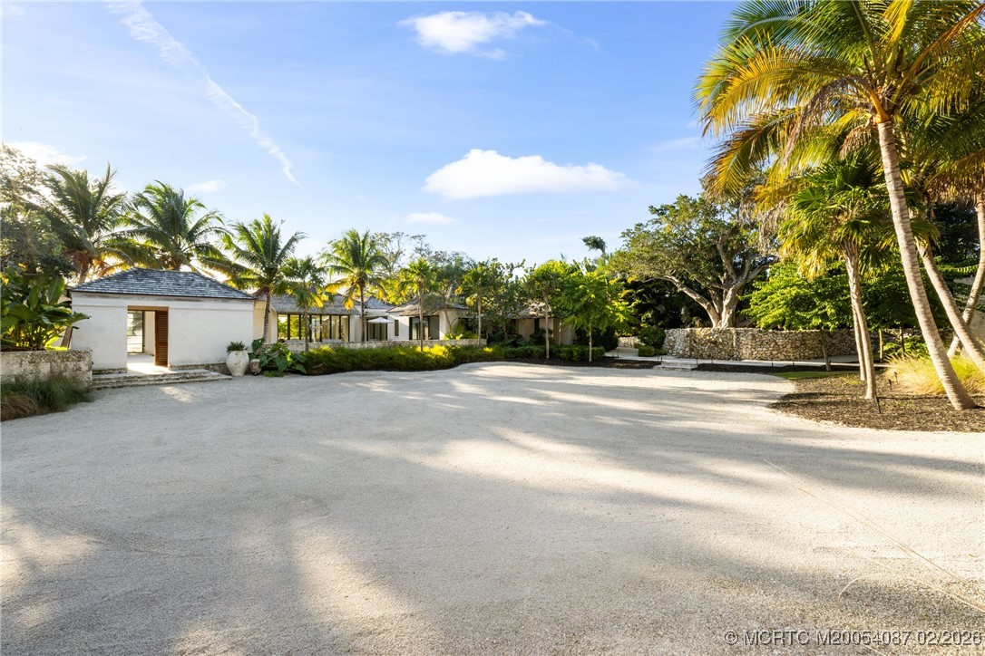 White home behind a broad driveway, framed by palm trees and lush landscaping.