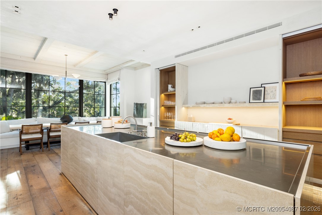 Modern kitchen with a large stone island, stainless-steel counter, warm wood cabinetry, and windows overlooking trees.