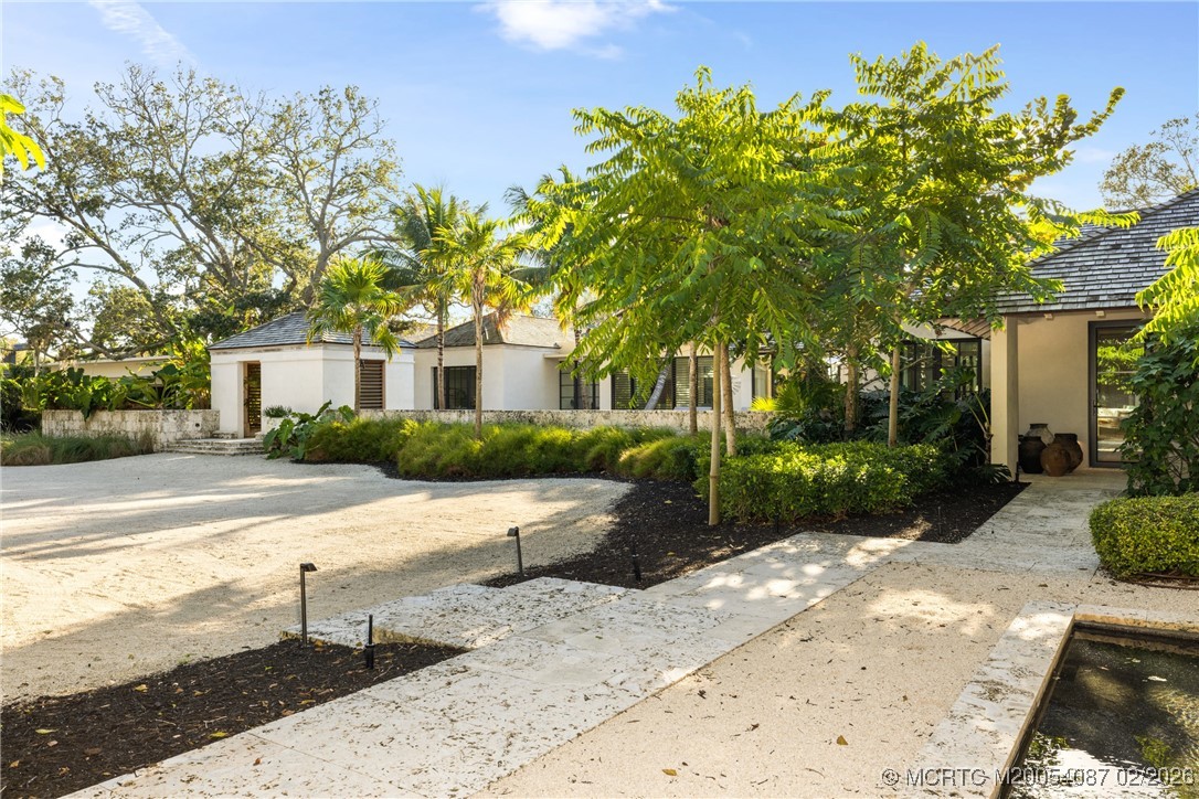 Stucco home behind tropical landscaping, with a gravel driveway, stone walkway, and courtyard water feature.