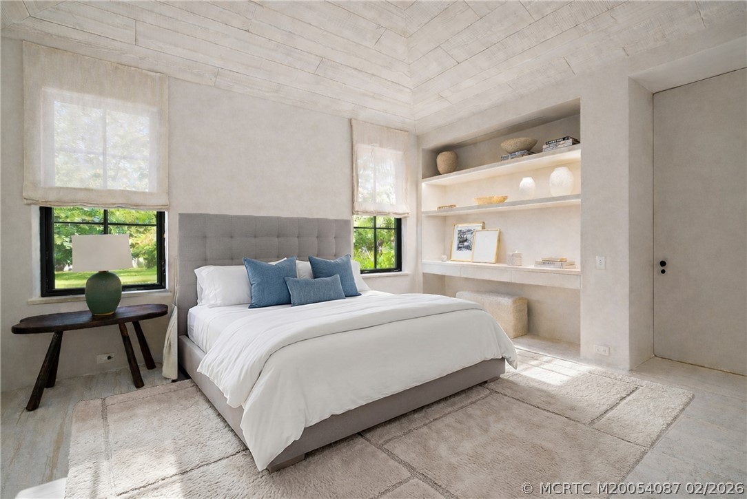 Bedroom with a gray upholstered bed, blue pillows, built-in display shelves, and windows overlooking greenery.
