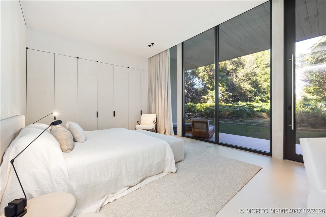 Modern bedroom with a white bed and floor-to-ceiling glass doors overlooking a landscaped garden.